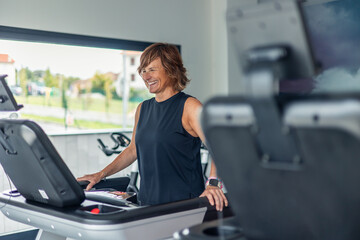 Fototapeta premium Smiling mature sporty woman walking on treadmill in gym for cardiovascular health