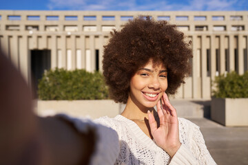 A woman with curly hair smiles and takes a selfie. She is wearing a white sweater