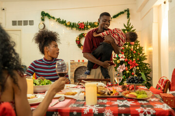 A joyful family gathers around the dinner table celebrating Christmas. The father, dressed in a Santa hat, gives a gift to the child, while everyone smiles with warmth and festive decorations.