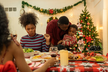 A joyful family gathers around the dinner table celebrating Christmas. The father, dressed in a Santa hat, gives a gift to the child, while everyone smiles with warmth and festive decorations.