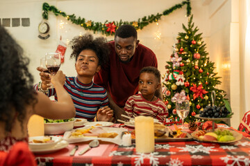 A joyful family gathers around the dinner table celebrating Christmas. The father, dressed in a Santa hat, gives a gift to the child, while everyone smiles with warmth and festive decorations.
