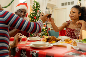 A joyful family gathers around the dinner table celebrating Christmas. The father, dressed in a Santa hat, gives a gift to the child, while everyone smiles with warmth and festive decorations.