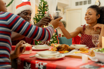 A joyful family gathers around the dinner table celebrating Christmas. The father, dressed in a Santa hat, gives a gift to the child, while everyone smiles with warmth and festive decorations.