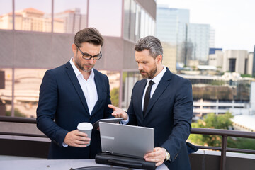 Two entrepreneurs near the office on the street. Business meeting of friends outdoors. Business mens with takeaway coffee and laptop outdoor. Business men team using laptop outdoor. Two businessman