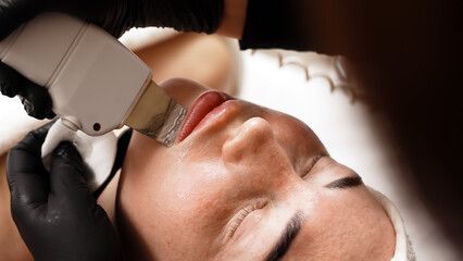 Close-up of a cosmetologist using an ultrasonic spatula for facial treatment on a young woman. The...