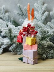 Christmas character rabbit with colorful ears in a sweater with presents in his hands on the background of pine branches