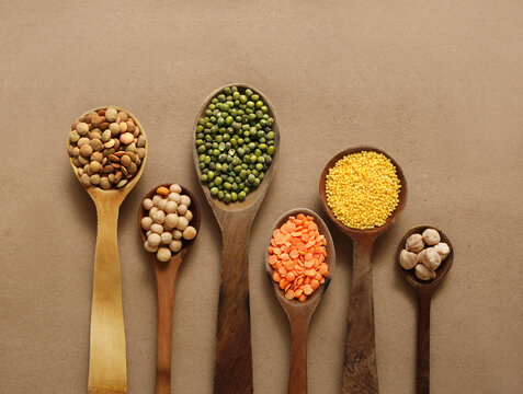 Wooden spoons with porridge, peas, dry millet groats, chickpeas, red lentil, mung beans on the brown background.
