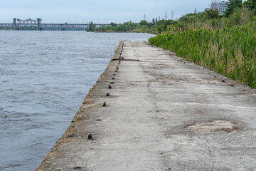 Support slabs along picturesque narrow river canal. Iron concrete reinforced pier from one bank of water to other. Trees bushes and reed grow along empty pedestrian. Reflection nature in river.