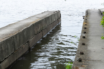 Fototapeta premium Support slabs along picturesque narrow river canal. Iron concrete reinforced pier from one bank of water to other. Trees bushes and reed grow along empty pedestrian. Reflection nature in river.