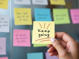 Close-up of a hand holding a sticky note with "Keep going" written, motivational message in an office setting, colorful sticky notes background, focus on persistence and continual effort towards goals