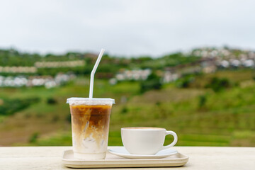 White cup of hot latte art coffee and iced latte coffee on cream wooden table with terrace mountain background in rainy season.