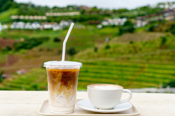 White cup of hot latte art coffee and iced latte coffee on cream wooden table with terrace mountain background in rainy season.