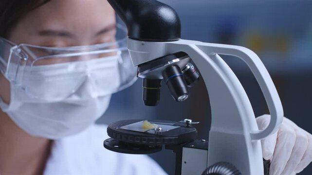 Close up video in high quality, a female researcher with safety goggles working in the clinical lab, adjusting microscope to look and analyze experiment sample. Copy space for text