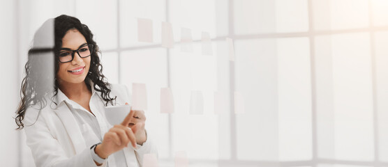 Paper Reminder. Smiling mexican businesswoman checking sticky notes glued to glass wall, drinking coffee at office