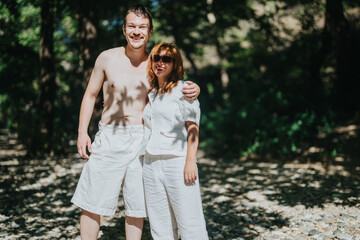 A cheerful couple poses in casual attire, embracing under the warm sunlight in a forest setting,...