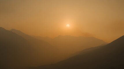 Obraz premium A golden sun sets behind rugged mountain silhouettes, casting a warm glow in the hazy atmosphere of late evening. The landscape is filled with soft shadows and tranquil colors as dusk approaches.