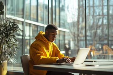 Young professional working on laptop, focused on task, sitting in modern office, bright yellow jacket, large glass windows, natural light, work environment concept