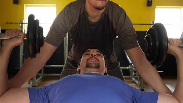 Man Bench Pressing Dumbbells with Personal Trainer Assistance. Man performing a chest workout with dumbbells while receiving help from a personal trainer, perfect for strength training.