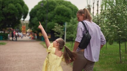 Seen from behind, mother and her young daughter with Down syndrome approach amusement park. Girl in yellow dress points excitedly while holding her mother's hand. Camera follows them