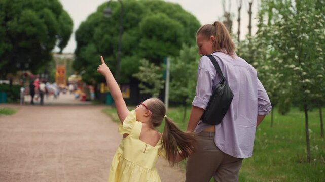 Seen from behind, mother and her young daughter with Down syndrome approach amusement park. Girl in yellow dress points excitedly while holding her mother's hand. Camera follows them