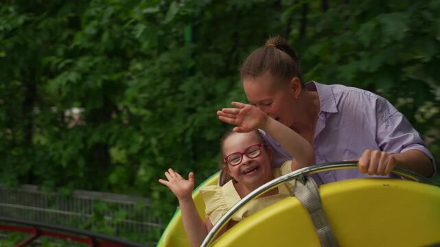 Young girl with Down syndrome and her mother are on a ride at amusement park. Girl has raised hands in excitement, and their cheerful expressions highlight delight of raising child with special needs