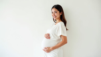 Obraz premium Young pregnant woman in a simple white outfit, smiling warmly and cradling her belly, against a plain white background.