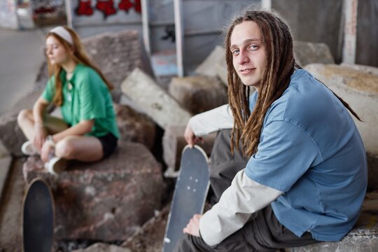 Portrait of young man with dreadlocks holding skateboard while sitting on rocks in urban area. Woman in background sitting with legs crossed on rocks - Powered by Adobe