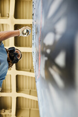 Artist wearing gas mask painting mural on wall under overpass, seen from below while spray can in...