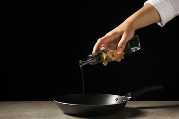 Woman pouring oil into frying pan at gray textured table against black background, closeup. Space for text