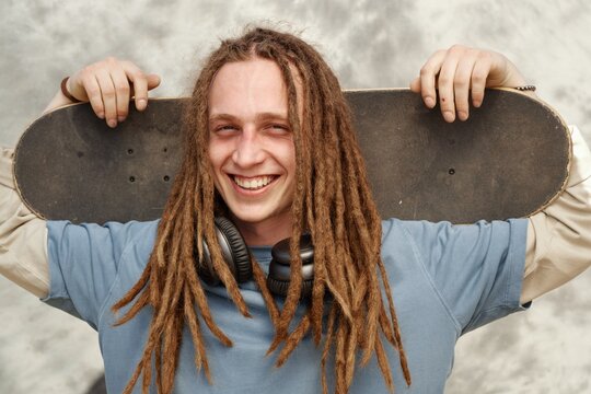 Caucasian man with long dreadlocks holding skateboard behind shoulders while smiling confidently with headphones around neck under chin and gray backdrop