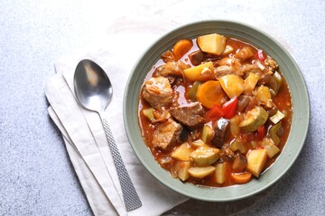 Delicious stew with vegetables in bowl and spoon on light grey table, top view