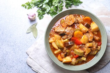 Delicious stew with vegetables in bowl, parsley, garlic and bay leaves on light grey table, top view