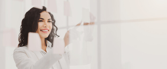 Sharing Ideas. Happy mexican businesswoman sticking adhesive notes on glass wall in office, copyspace
