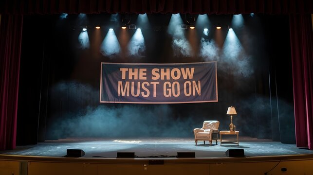 An empty stage with spotlights and a smoke banner in the background. The stage has a chair and a lamp. The smoke banner has the text "The show must go on".