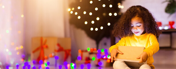 Little african american girl opening magic gift box, sitting near Christmas tree, free space
