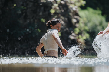 Fototapeta premium A happy woman joyfully splashes water in a beautiful natural setting, surrounded by lush greenery. The image captures the essence of fun, summer, and outdoor adventure.