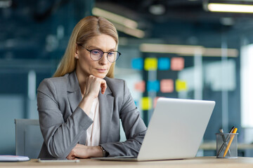 Confident businesswoman using laptop in office. She is deep in thought while analyzing data for...