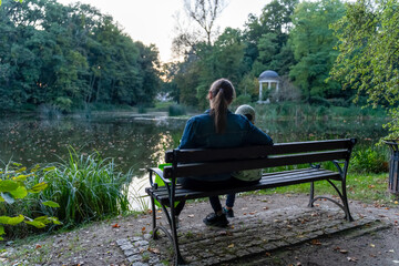 Mother and child sitting on a bench by a quiet lake in a park at dusk. Concept of quality family holidays, tranquility and unity with nature. High quality photo