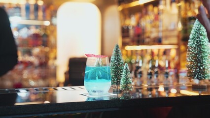 Bartender serving Christmas blue Lagoon cocktail with red dried fruit rosemary design on luxury bar counter with VIP group guest friend or couple in green neon snow Christmas tree festive. Vinosity.