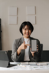 Confident Businesswoman Presenting Smartphone: A young Asian businesswoman in a professional setting, confidently pointing to a blank smartphone screen with a bright smile, conveying a sense of profes