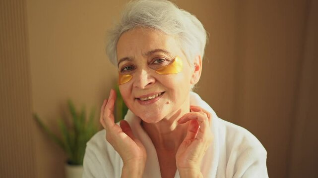 Eye patch beauty aging treatment. Mature adult old woman with under eye collagen pads. Senior woman with patches under eyes gets facial treatment. Senior lady in robe relaxing in bathroom at home