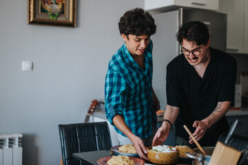 Two young men are joyfully preparing a pasta meal at home. They are standing around a table with plates and a bowl of salad, enjoying their cooking time together.