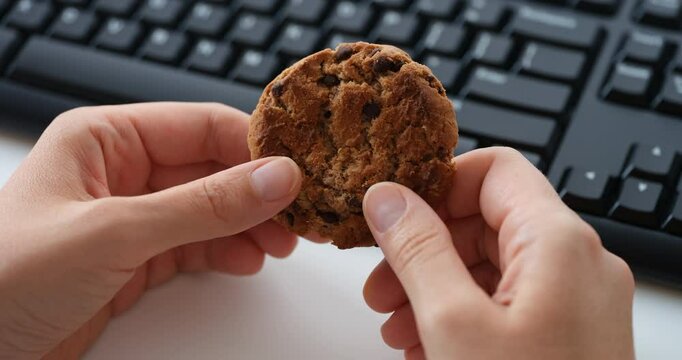 Woman holding a chocolate chip cookie in her hands above a computer keyboard and getting ready to eat it during a break at work. Close up.