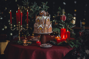 Elegant luxury Christmas table setting for family home party with homemade big gingerbread house, holiday celebration. Red tablecloth, candles, chandelier. Romantic candlelit dinner