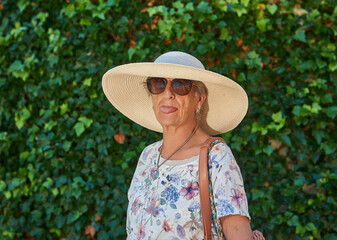 An octogenarian woman wearing sunglasses, a sun hat and a shoulder bag sticks out her tongue mockingly as she sunbathes in front of an ivy-covered wall