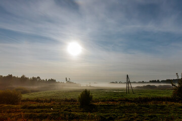 A serene field bathed in soft sunlight, with a light mist hovering over the grass. Power lines and poles stretch across the landscape, and trees line the horizon. The atmosphere is peaceful