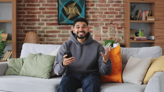 Front view of handsome hindu man screaming ecstatically while looking at camera with mobile phone in hand. Cheerful person in grey hoodie with beard sitting on sofa in flat with stylish interior.