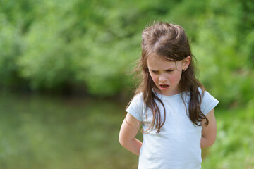 Little cute preschool girl in a T-shirt plays on the background of a pond. Happy childhood concept outdoors