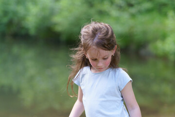 Little cute preschool girl in a T-shirt plays on the background of a pond. Happy childhood concept outdoors