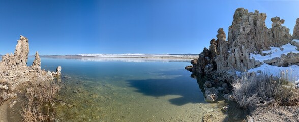 This panoramic image showcases the striking salt rock formations at Mono Lake, California. The crystal-clear, still waters of the lake reflect the vivid blue sky, creating a serene atmosphere.